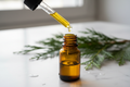 oil dropper dropping essential oil into an amber colored bottle with cypress herb in the background